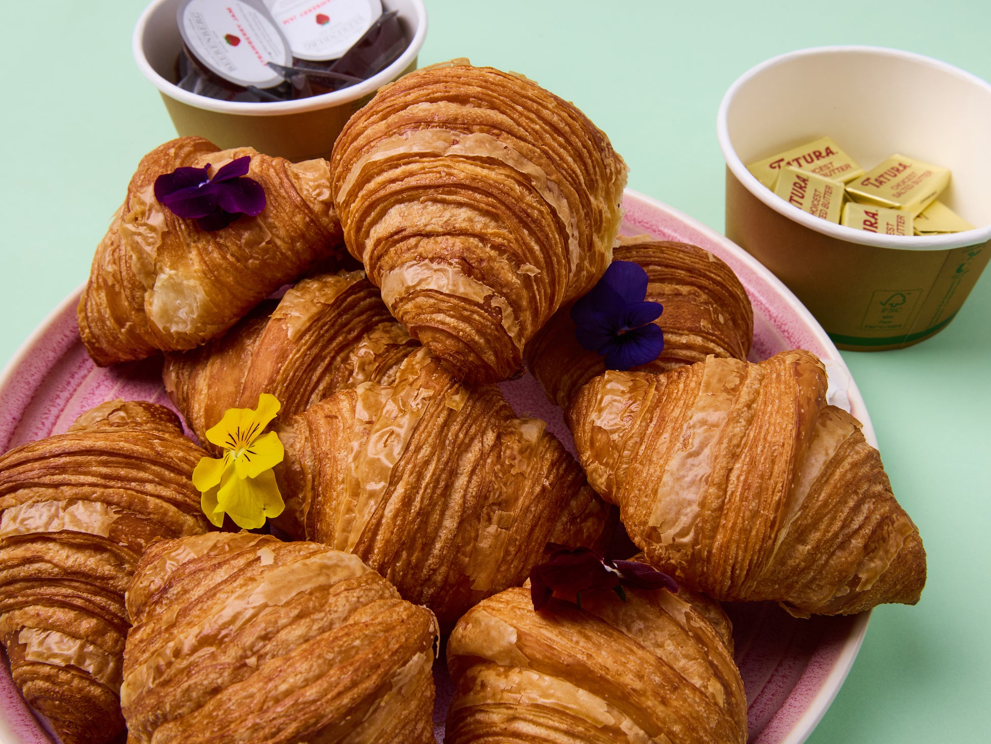 Croissants on a pink plate with small flowers and cups of butter and jam in the background.